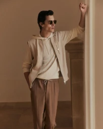 Model Liam Kelly wearing a cream hooded sweater, beige zip vest, and camel trousers leaning against an architectural stone arch.