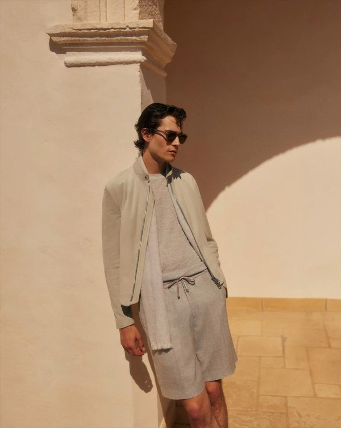 Liam Kelly wears a neutral zip jacket, knit top, and drawstring shorts with sunglasses while leaning against a sunlit stone wall.