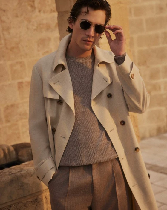 Male model wearing a cream trench coat adjusting sunglasses against a stone building exterior.