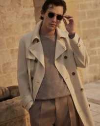Male model wearing a cream trench coat adjusting sunglasses against a stone building exterior.