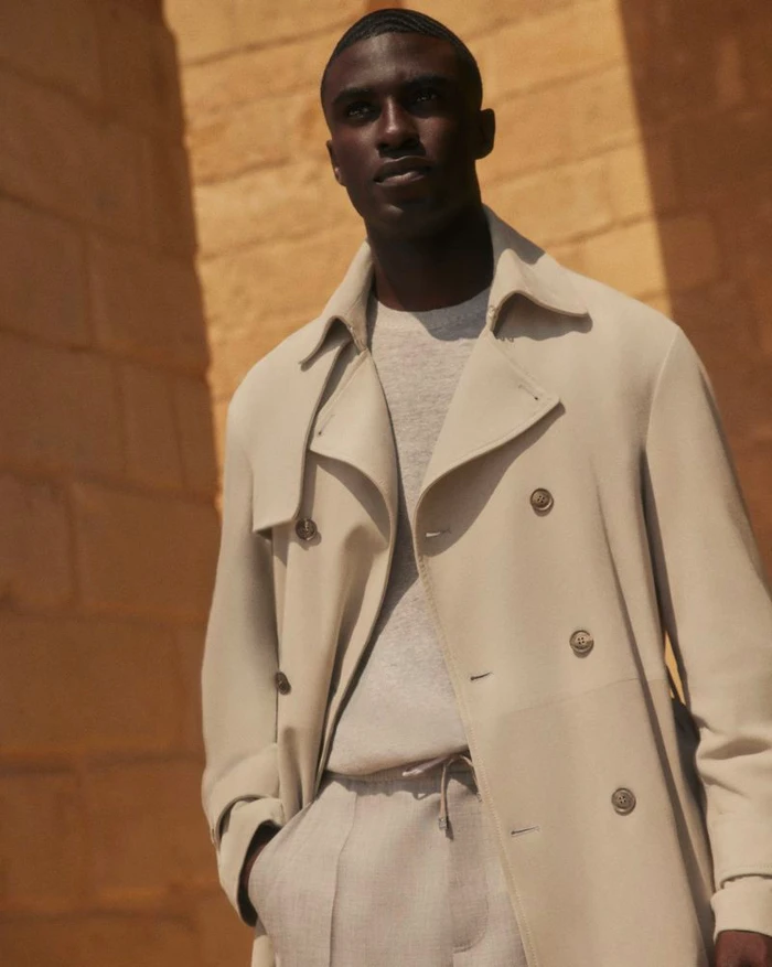 Model wearing a light beige double-breasted trench coat outdoors against stone architecture for Brett Johnson.