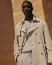 Model wearing a light beige double-breasted trench coat outdoors against stone architecture for Brett Johnson.