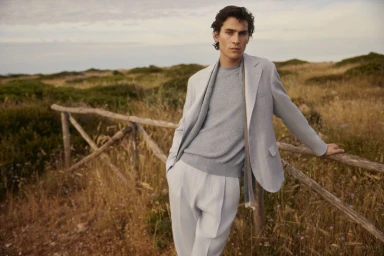 Model leaning on a wooden fence wearing a light grey suit jacket and matching trousers in a grassy rural landscape.