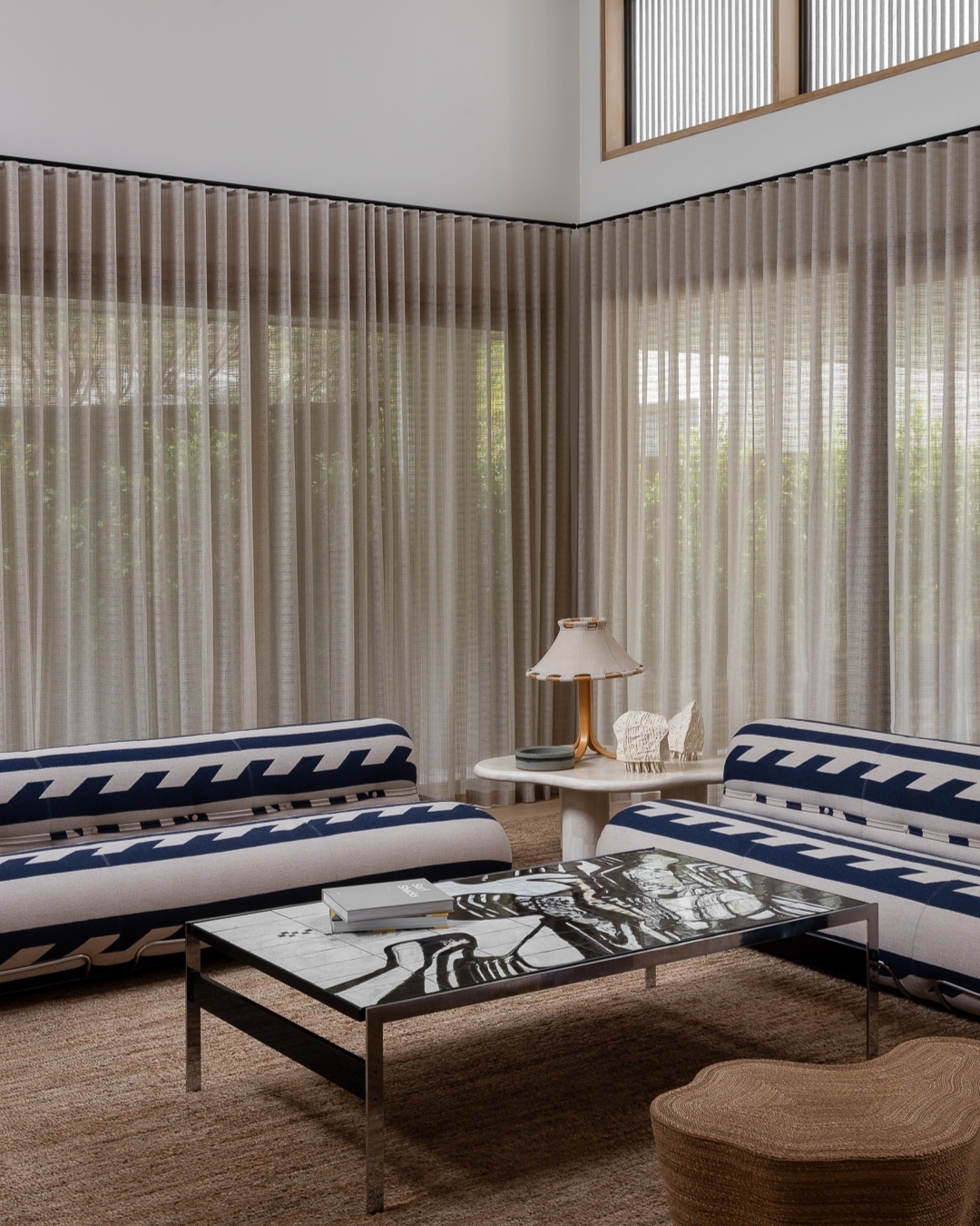 Living room interior featuring a sofa, patterned coffee table, and natural light filtering through sheer curtains in a neutral color scheme.
