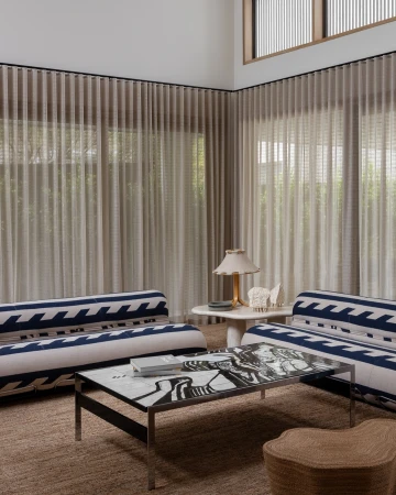 Living room interior featuring a sofa, patterned coffee table, and natural light filtering through sheer curtains in a neutral color scheme.