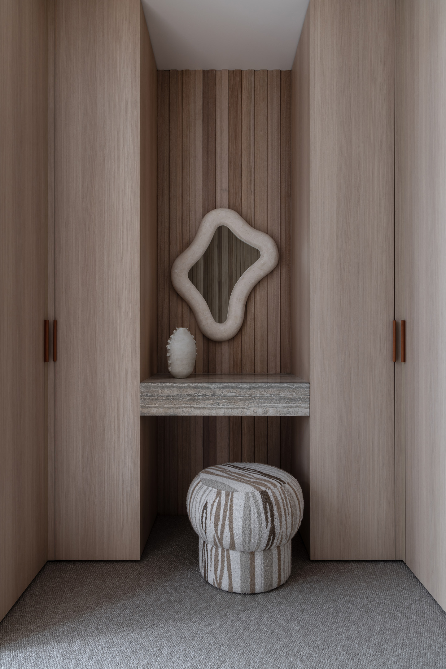 Modern dressing area with a sculptural mirror, vanity, and ottoman, framed by symmetrical light wood wardrobes, creating a serene, stylish interior.