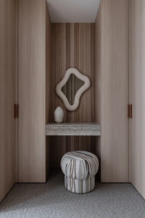 Modern dressing area with a sculptural mirror, vanity, and ottoman, framed by symmetrical light wood wardrobes, creating a serene, stylish interior.