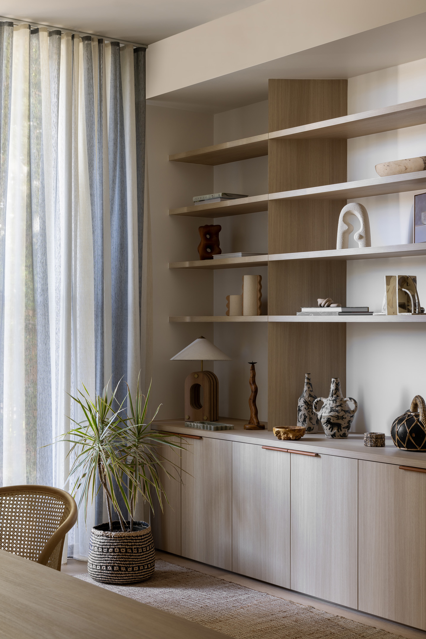 Interior view of a living space with wood-paneled cabinets, shelves displaying decorative objects, a potted plant, and sheer striped curtains.