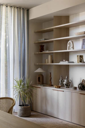 Interior view of a living space with wood-paneled cabinets, shelves displaying decorative objects, a potted plant, and sheer striped curtains.