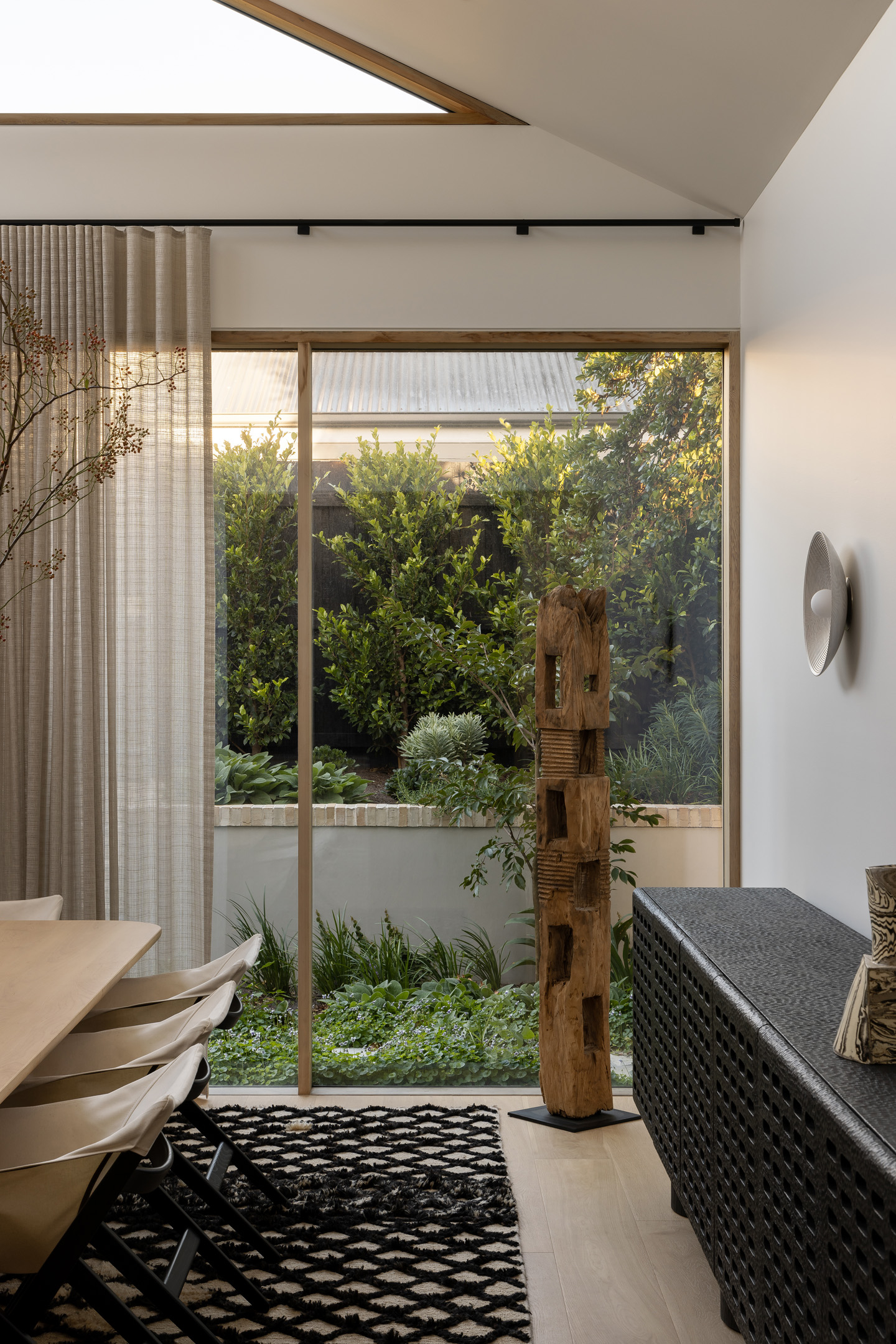 Stylish interior features a dining table with chairs, wooden sculpture, and textured sideboard, set against a green outdoor garden backdrop.