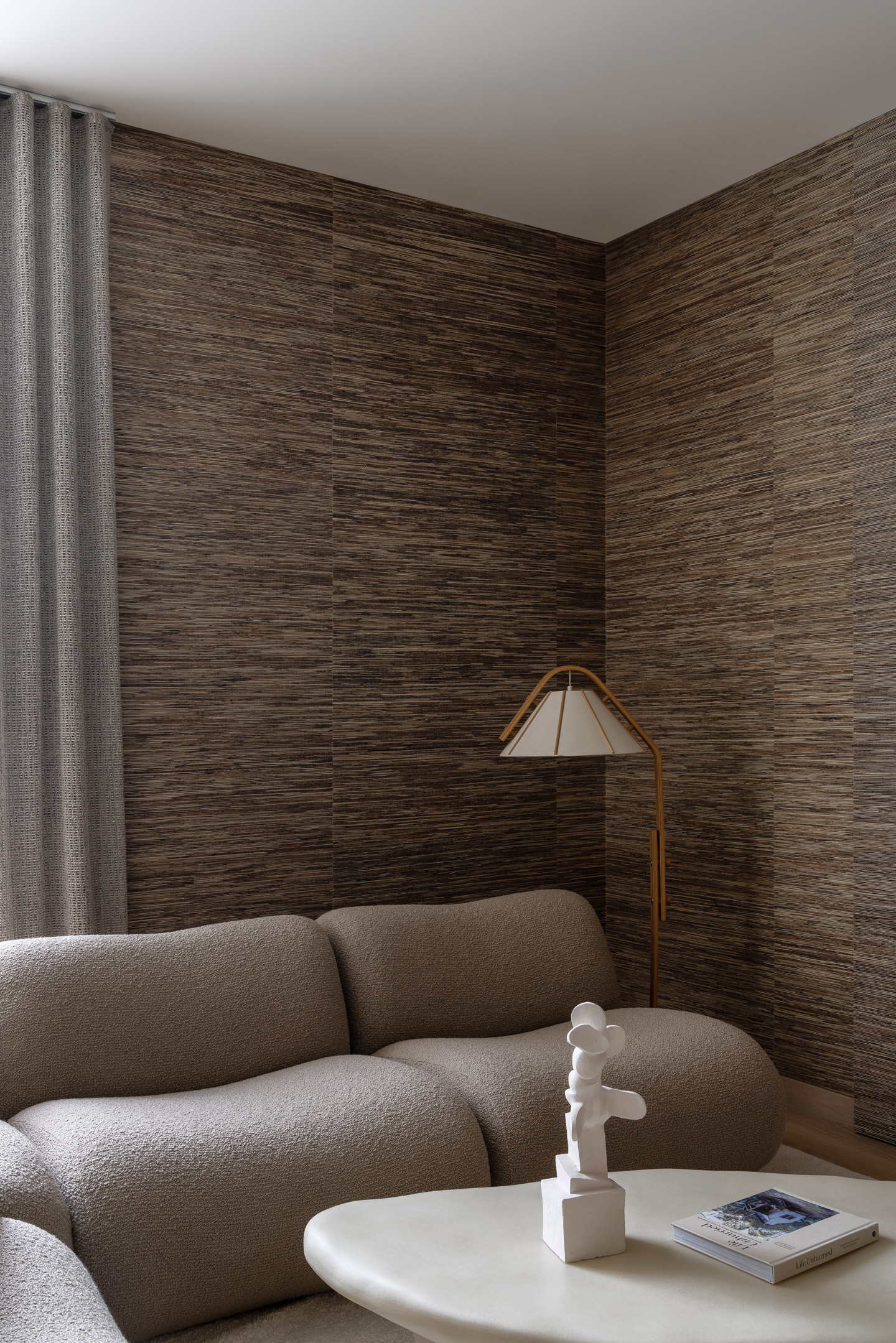 Interior corner with light brown textured wallpaper, a beige rounded sofa, a floor lamp, and a modern white sculpture on a coffee table.