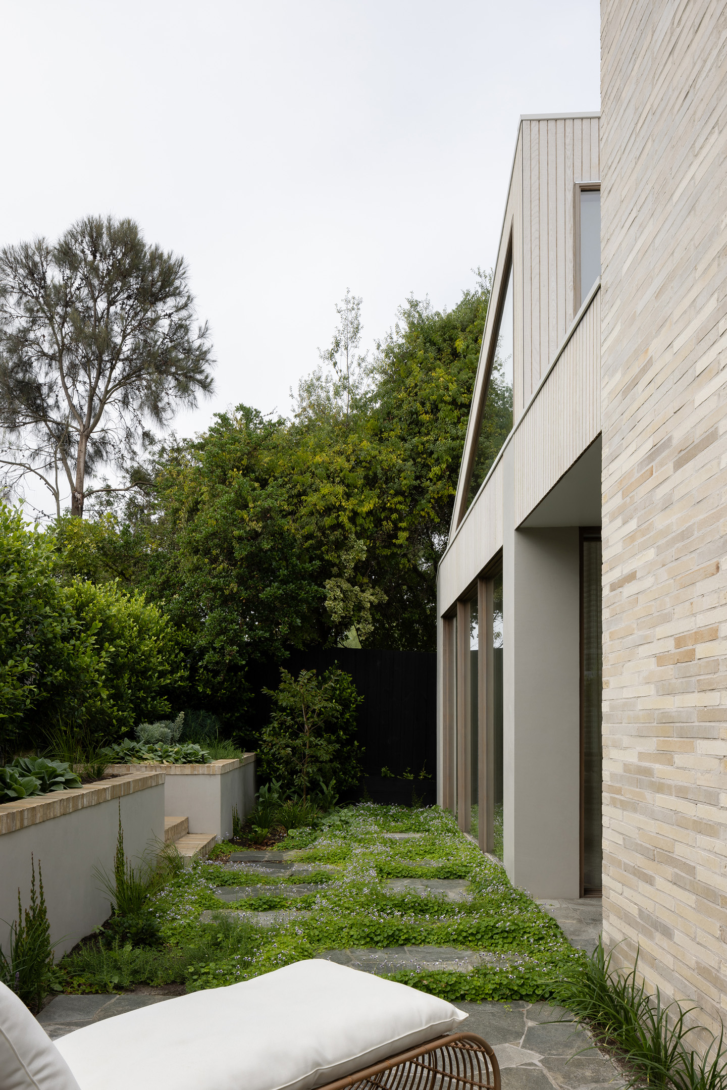 Exterior view of a modern, light brick home with large windows, surrounded by lush greenery and a manicured garden on an overcast day.