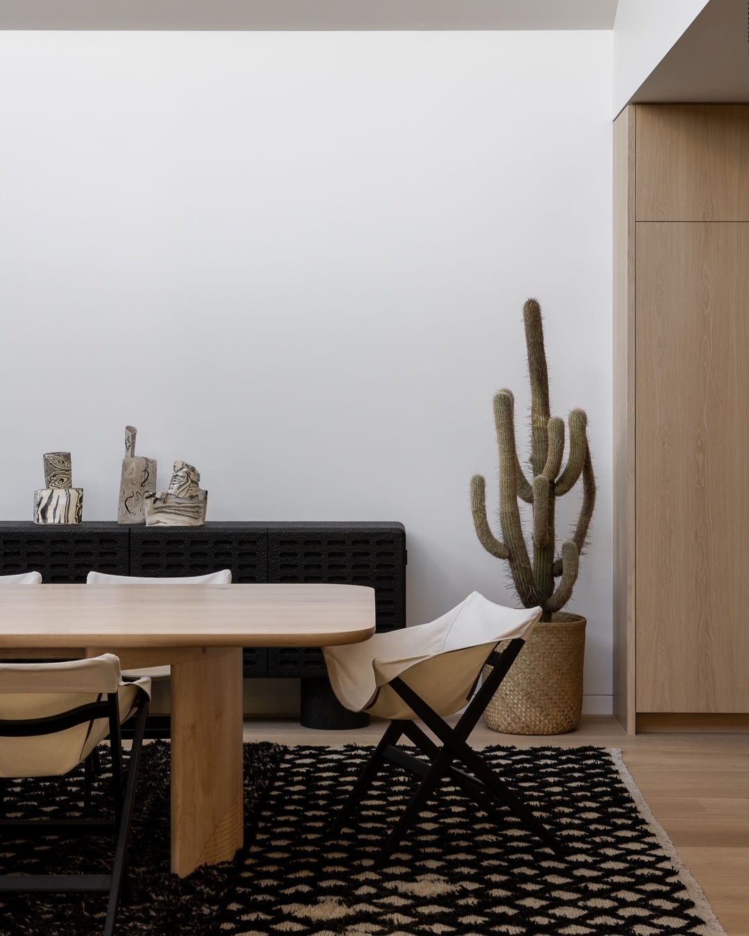 Minimalist interior featuring a light wood table, black accent chair, and a tall cactus against a white wall. Rug with geometric pattern.