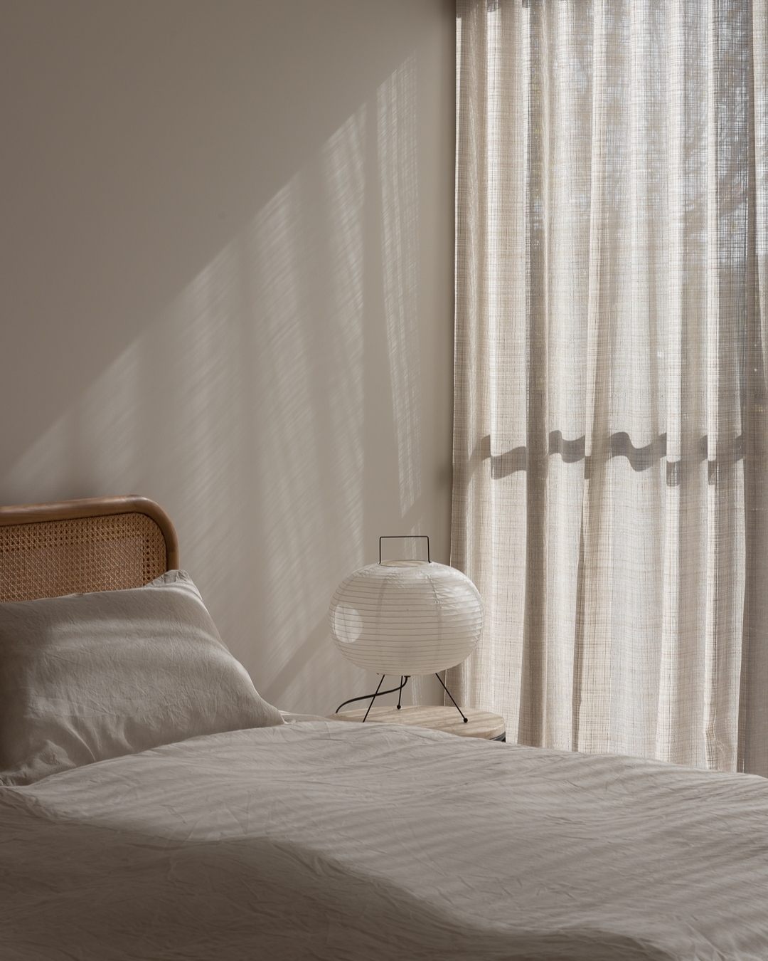 Bedroom interior featuring a linen-covered bed, a rattan headboard, a paper lantern on a bedside table, and sunlight filtering through curtains.