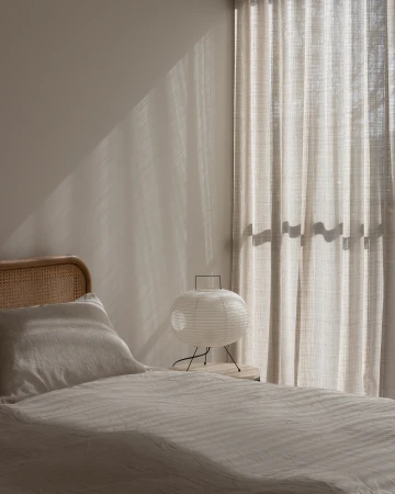 Bedroom interior featuring a linen-covered bed, a rattan headboard, a paper lantern on a bedside table, and sunlight filtering through curtains.