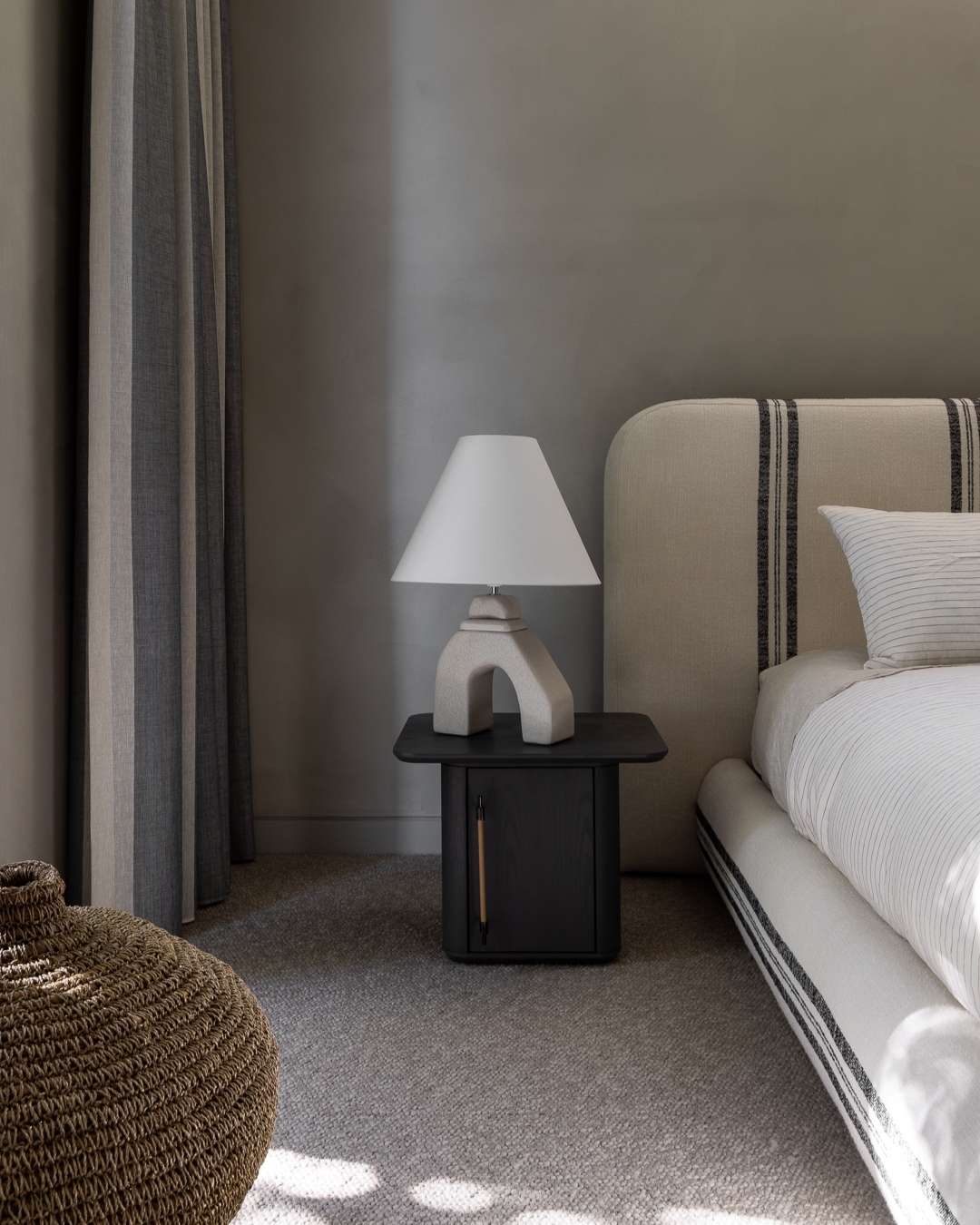 Bedroom interior featuring a striped bed, contemporary lamp on a black nightstand, woven basket, and gray wall with window-lit shadows.