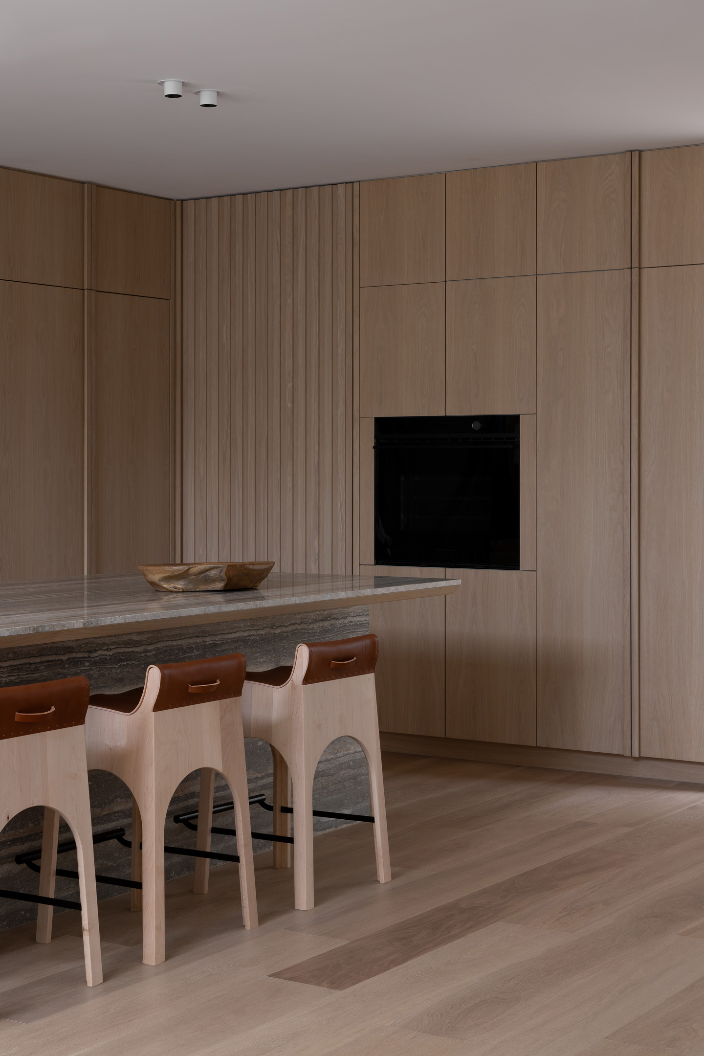 Modern kitchen interior featuring wooden cabinets, marble countertop island with stools, and a built-in black oven.