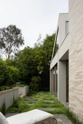 Exterior view of Barwon Heads Residence featuring light brick architecture, lush garden landscaping, and a lounge chair in the foreground.