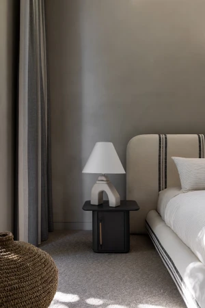 Modern bedroom corner featuring a small black bedside table, sculptural ceramic table lamp, and upholstered bed with striped headboard.