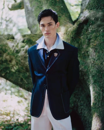 Model wearing a dark blazer and layered shirt posing outdoors against a large mossy tree trunk