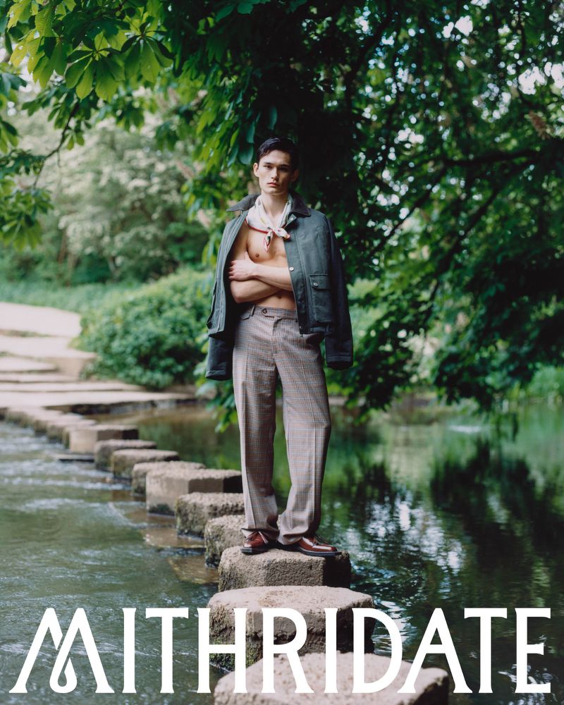 Model Alex Schlab poses shirtless with a dark jacket and tailored trousers on stone steps across a stream in lush greenery.