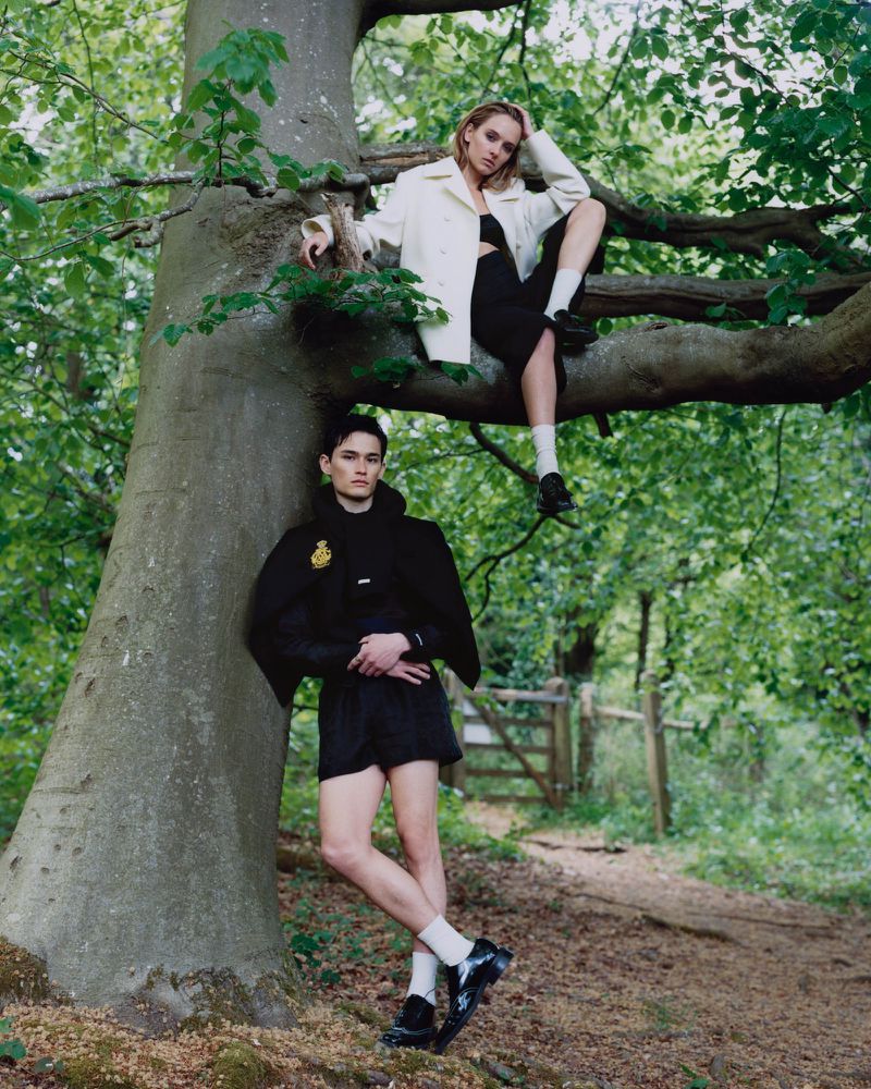 Two models posing outdoors in a wooded setting, one seated in a tree and one standing leaning against the trunk.