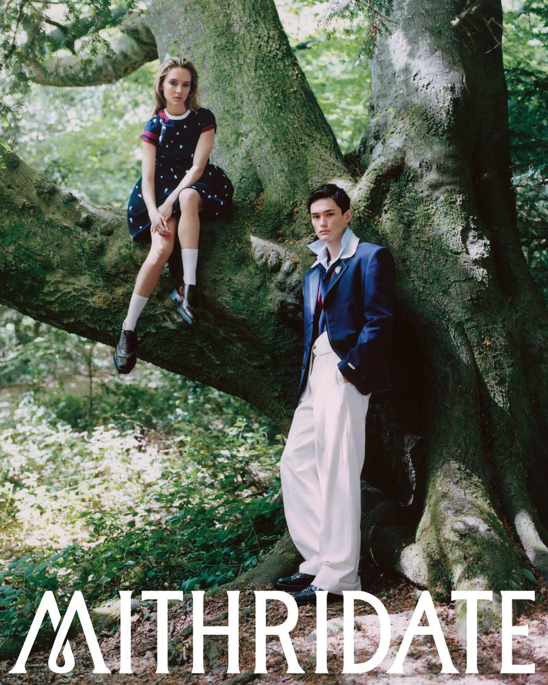 Two models posing in tailored blue and white fashion outfits beside a large moss-covered tree in a lush green forest.
