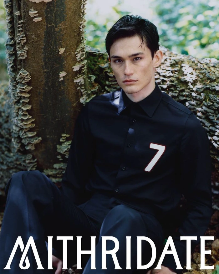Model Alex Schlab seated in an all-black collared shirt with a number seven patch against a textured mossy tree.