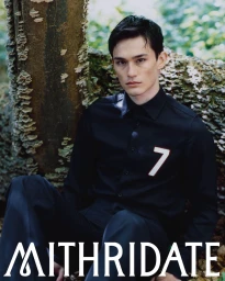 Model Alex Schlab seated in an all-black collared shirt with a number seven patch against a textured mossy tree.