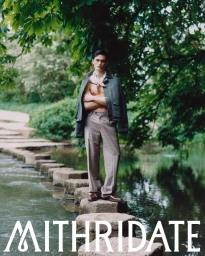 Model Alex Schlab poses shirtless with a dark jacket and tailored trousers on stone steps across a stream in lush greenery.