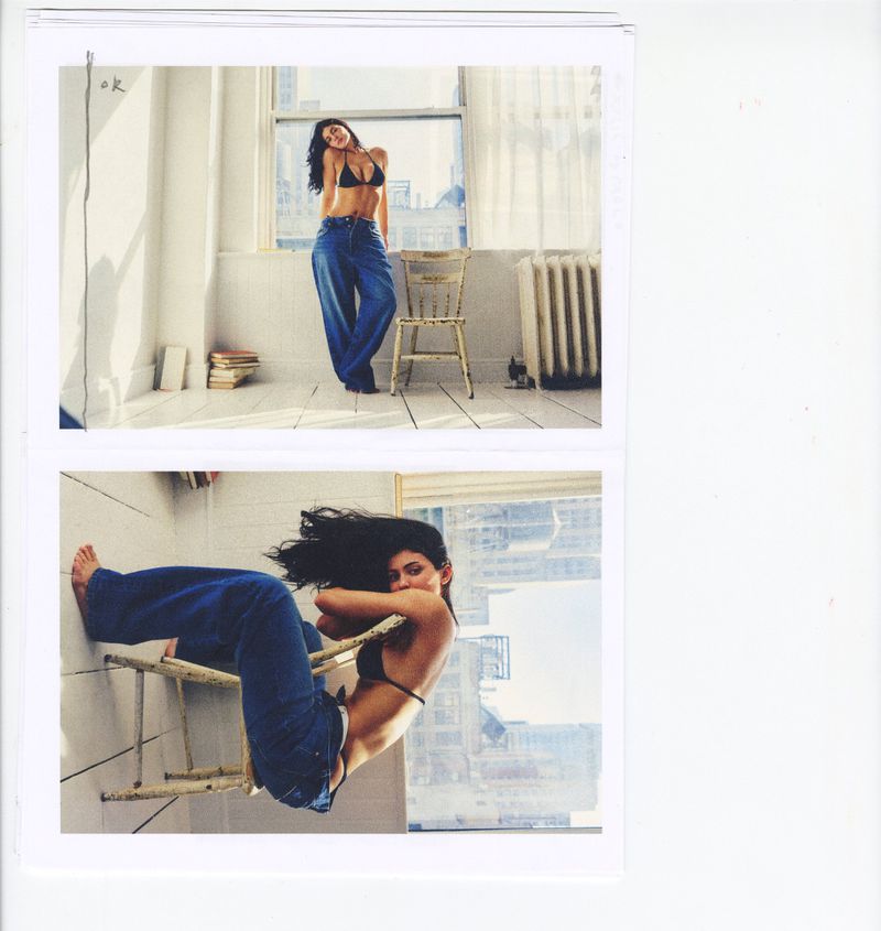 Kylie Jenner posing in a black bikini top and baggy blue denim jeans next to a rustic wooden chair by a bright window.