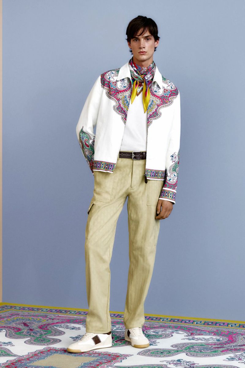 Male model in a white paisley-trimmed jacket, beige trousers, and patterned neck scarf standing on a matching carpet against a blue wall.