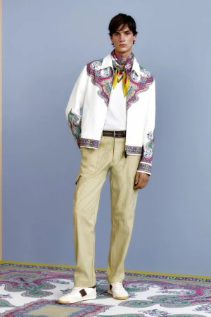 Male model in a white paisley-trimmed jacket, beige trousers, and patterned neck scarf standing on a matching carpet against a blue wall.