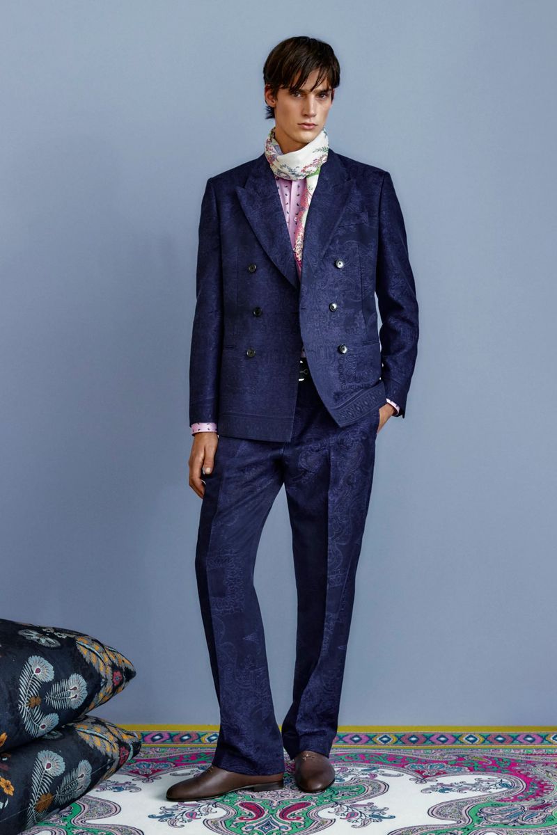 Male model in a navy patterned double-breasted suit and patterned scarf posing in front of a light blue wall, fashion editorial.