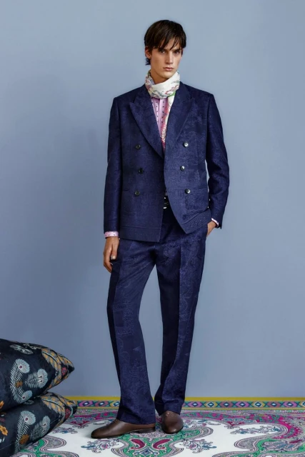 Male model in a navy patterned double-breasted suit and patterned scarf posing in front of a light blue wall, fashion editorial.