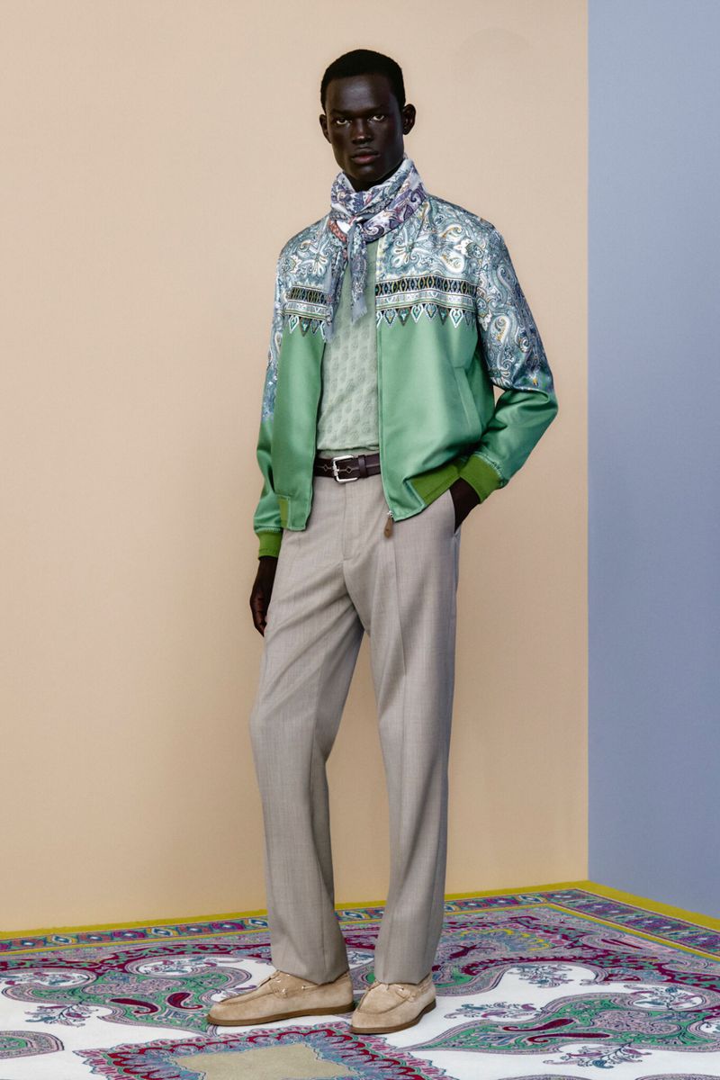 A man in a patterned jacket and scarf poses on a paisley rug in front of a two-toned beige and blue wall in a fashion editorial.