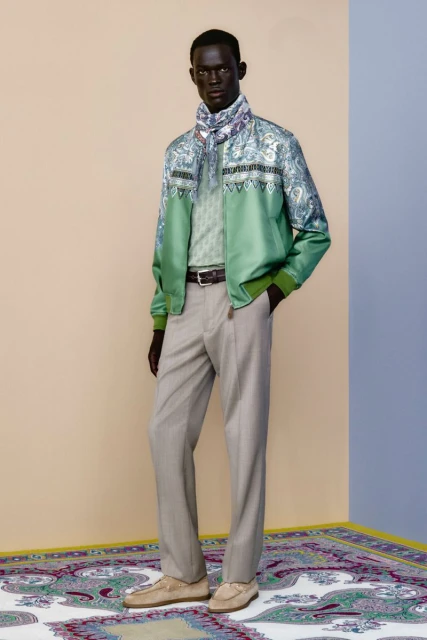 A man in a patterned jacket and scarf poses on a paisley rug in front of a two-toned beige and blue wall in a fashion editorial.