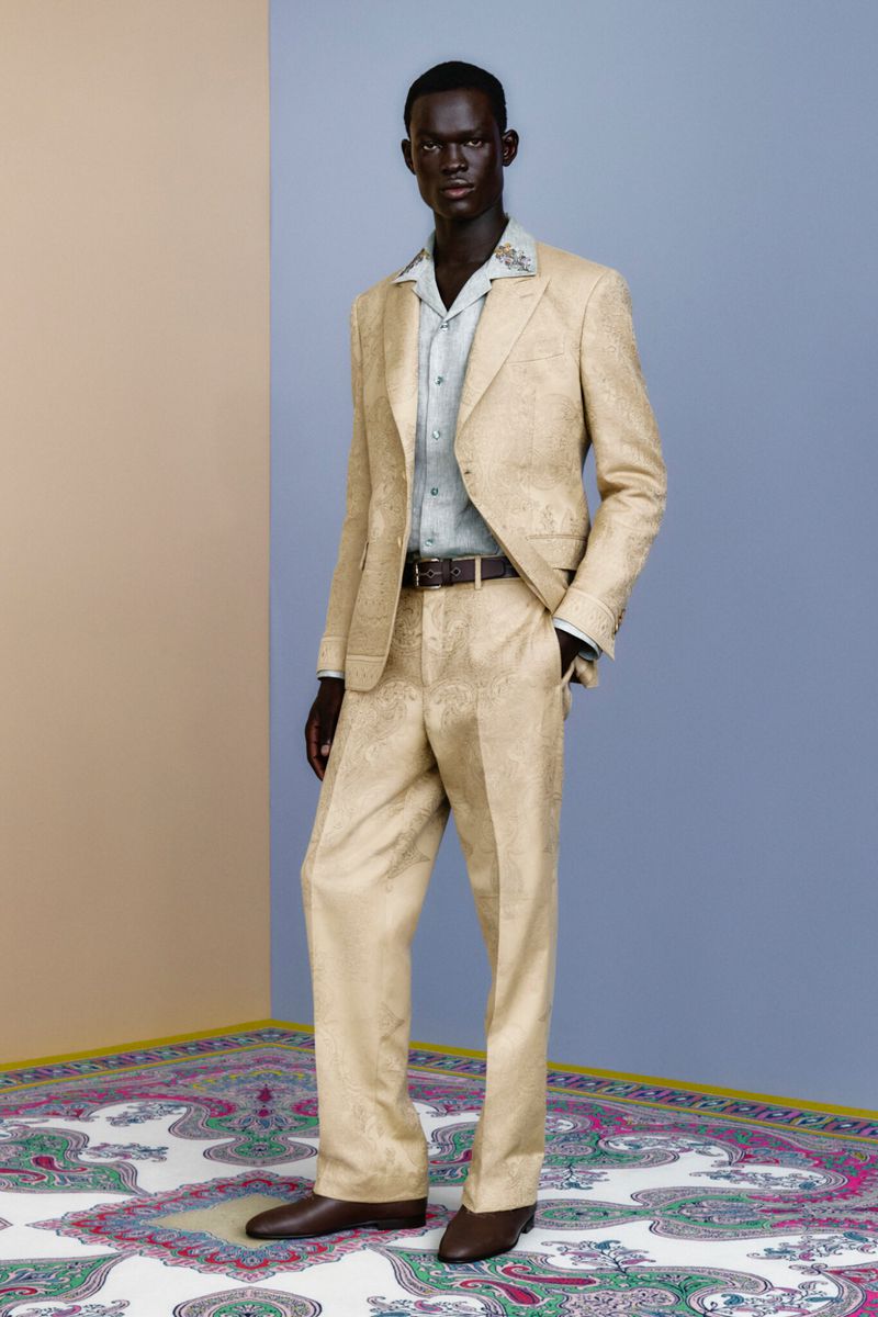 An adult male model poses in a tan patterned suit with a light-blue collared shirt and brown shoes in a minimalist photo studio.