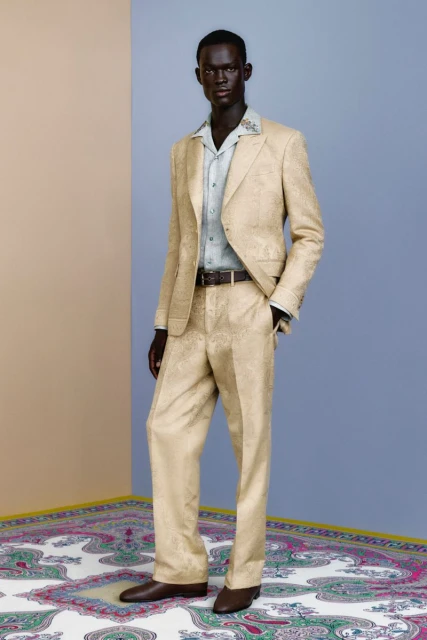 An adult male model poses in a tan patterned suit with a light-blue collared shirt and brown shoes in a minimalist photo studio.