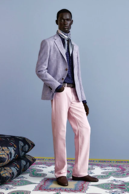 A Black male model poses in a light purple linen blazer, pink dress pants, and brown leather loafers against a pale blue backdrop.