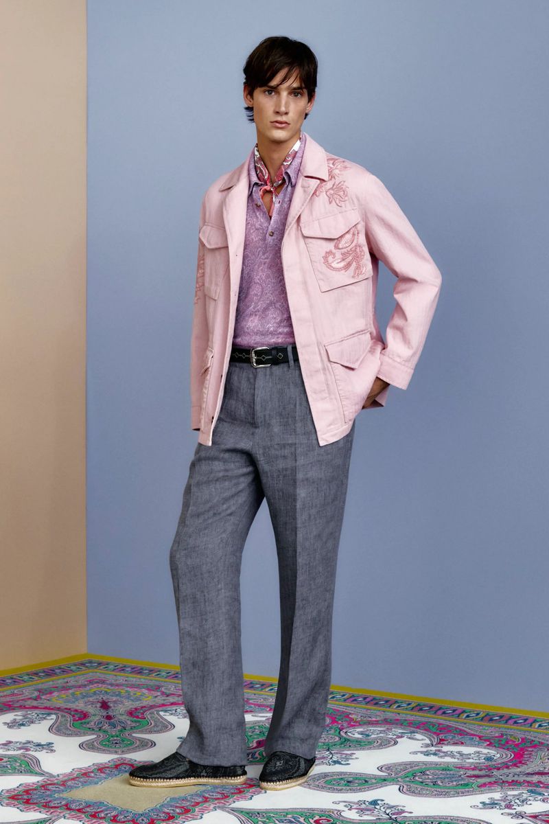 Male fashion model posing in a studio wearing a pink embroidered safari jacket, paisley shirt, grey trousers, and black loafers.