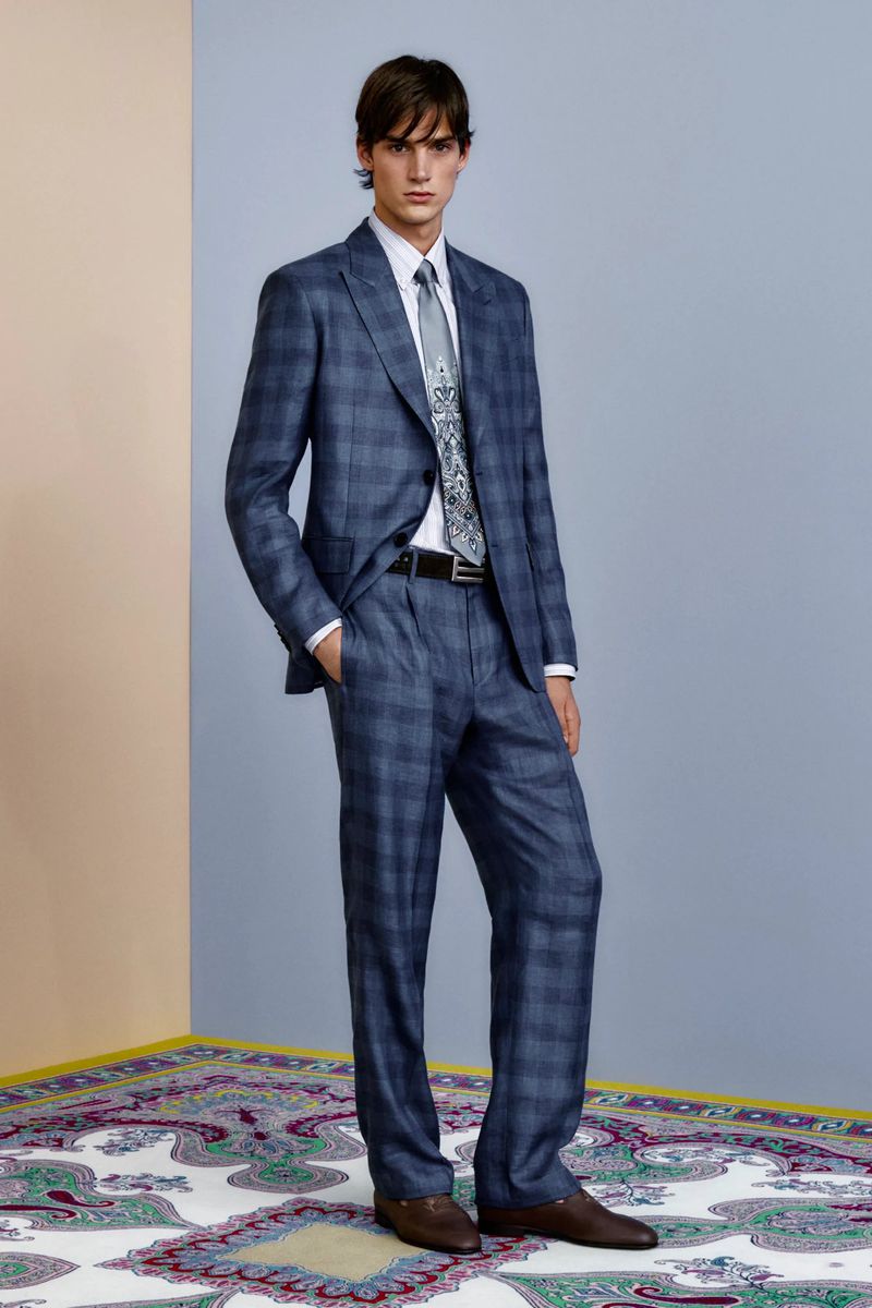 Fashionable adult male in a blue plaid suit with a patterned tie, standing in a studio with an ornate rug and blue wall.