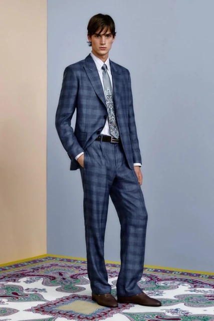 Fashionable adult male in a blue plaid suit with a patterned tie, standing in a studio with an ornate rug and blue wall.