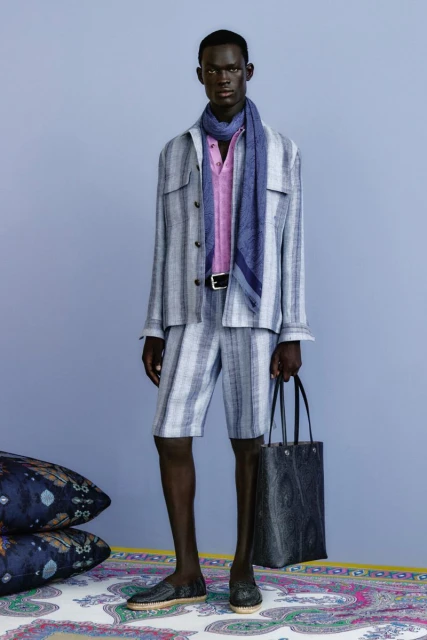 An African-American male model poses indoors wearing a striped linen suit with a pink linen shirt, blue scarf, black belt, and matching shoes.