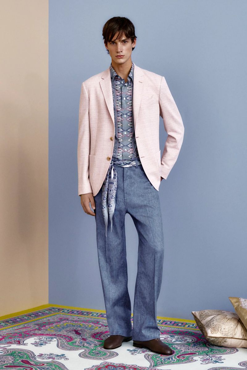 A stylish male fashion model in a pink blazer, patterned shirt, and blue trousers poses indoors against a two-tone wall.