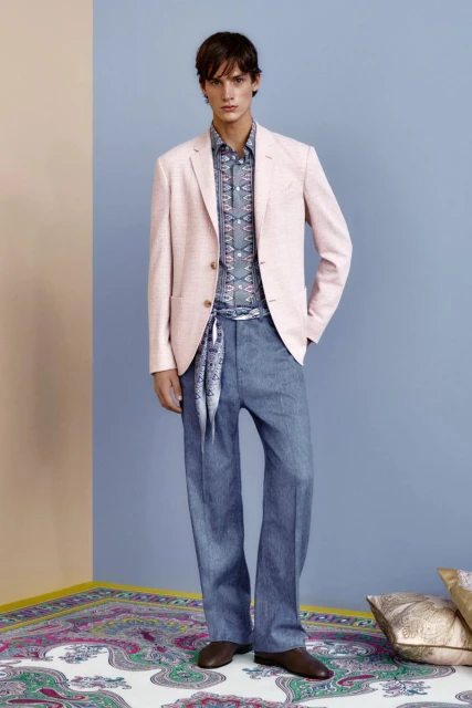 A stylish male fashion model in a pink blazer, patterned shirt, and blue trousers poses indoors against a two-tone wall.