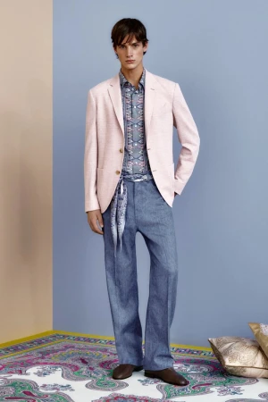A stylish male fashion model in a pink blazer, patterned shirt, and blue trousers poses indoors against a two-tone wall.