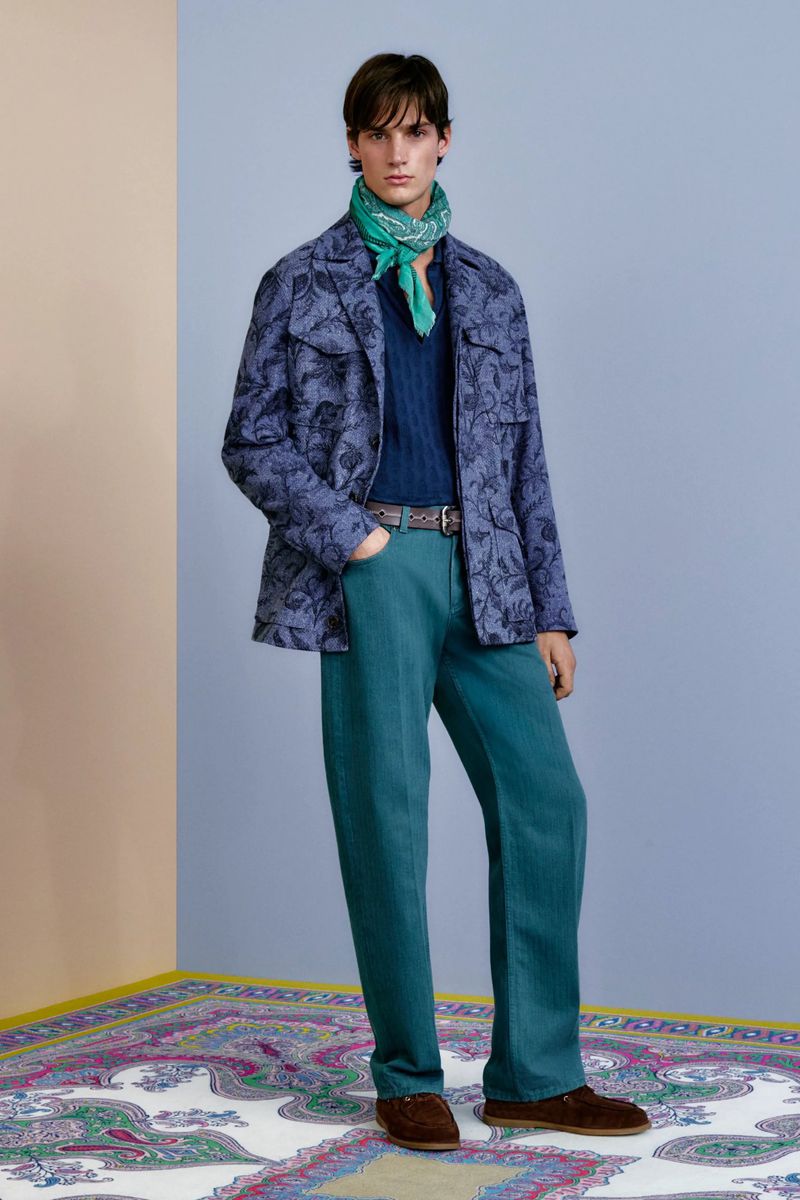 Full-length shot of a male model wearing a patterned jacket, navy top, teal trousers, brown shoes, and a green scarf in a studio.