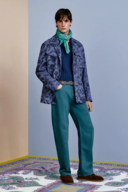 Full-length shot of a male model wearing a patterned jacket, navy top, teal trousers, brown shoes, and a green scarf in a studio.