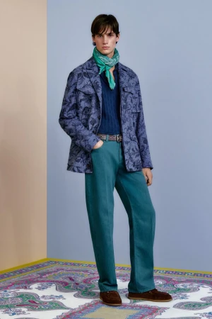 Full-length shot of a male model wearing a patterned jacket, navy top, teal trousers, brown shoes, and a green scarf in a studio.